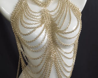 May include: Gold-colored chainmail halter top. The top is displayed on a white mannequin. The chainmail is made of small, interlocking metal rings that drape over the chest and shoulders. The design is intricate and decorative.