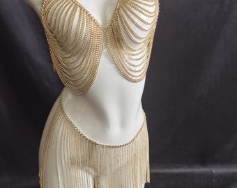 Può includere: Set top e gonna in cotta di maglia color oro. Il top è uno stile halter con catene drappeggiate e la gonna presenta un design a frange. Il set è esposto su un manichino bianco su sfondo scuro.