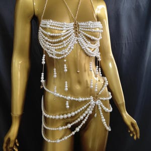 Könnte beinhalten: Eine goldene Schaufensterpuppe präsentiert eine Perlen-Körperkette. Das Oberteil ist im Neckholder-Stil mit mehreren Perlensträngen. Taille und Hüfte sind mit Perlensträngen und goldenen Kettenakzenten verziert. Drapierte Perlenstränge hängen von Oberteil und Taille. Der Hintergrund ist dunkel.