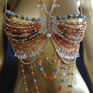 May include: A beaded top with a gold chain strap and a gold chain back. The top is made of orange, blue, and clear beads. The beads are arranged in rows and connected with gold chain. The top is displayed on a gold mannequin.