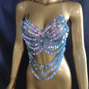 May include: A halter-style crop top constructed from silver-toned metal chains and iridescent, light-reflecting sequins. The garment features multiple draped layers of circular, shell-like discs in shades of blue, pink, and purple, creating a shimmering effect.