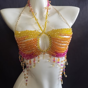 Puede incluir: Top halter con cuentas en tonos amarillo, naranja y rosa. La parte superior está hecha de pequeñas cuentas brillantes dispuestas en filas horizontales. Una cadena dorada forma las correas del halter. Los detalles de cuentas colgantes añaden movimiento y estilo.