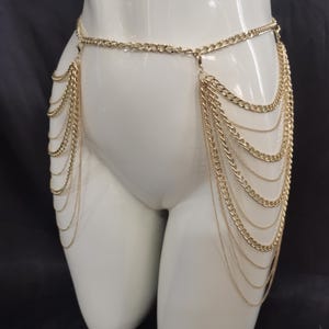 Könnte beinhalten: Goldfarbene Körperkette mit mehreren Strängen zarter Ketten, die von der Taille herabfallen. Die Kette ist über einer Schaufensterpuppe drapiert, um das Design und die Details des Schmucks zu zeigen. Ein stilvolles Accessoire.