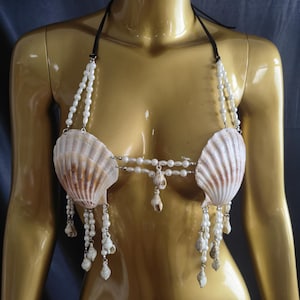 May include: A seashell bikini top with a black halter neck. The cups are made of large, cream-coloured seashells, adorned with white pearl beads and small dangling shells. The design evokes a mermaid or beach theme.