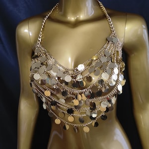 May include: A gold-colored halter top made of chains and circular sequins. The top is designed to cover the chest and is displayed on a mannequin. The sequins are arranged in multiple layers, creating a shimmering effect. The background is a dark blue.