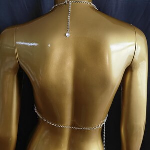 May include: A silver-toned body chain featuring a delicate link construction that wraps around the torso. The piece includes a necklace-style chain around the neck with a small circular charm at the end of the adjustable extender chain.