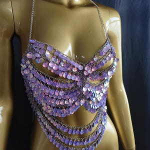 May include: A halter-style body chain top constructed from silver-toned metal links and adorned with numerous iridescent, shell-shaped sequins that shimmer with purple and pink hues. The garment features multiple draped layers of these reflective discs.