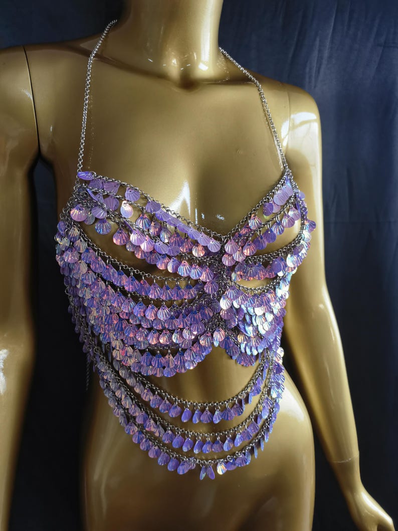 May include: A halter-style body chain top constructed from silver-toned metal links and adorned with numerous iridescent, shell-shaped sequins that shimmer with purple and pink hues. The garment features multiple draped layers of these reflective discs.