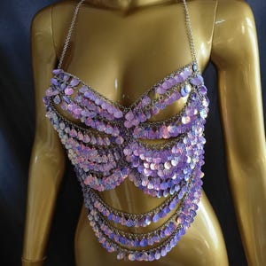 May include: A halter-style top constructed from silver-toned metal chains and iridescent, light purple, shell-shaped sequins. The garment features multiple draped layers of these shimmering, scale-like discs that create a textured, reflective surface.