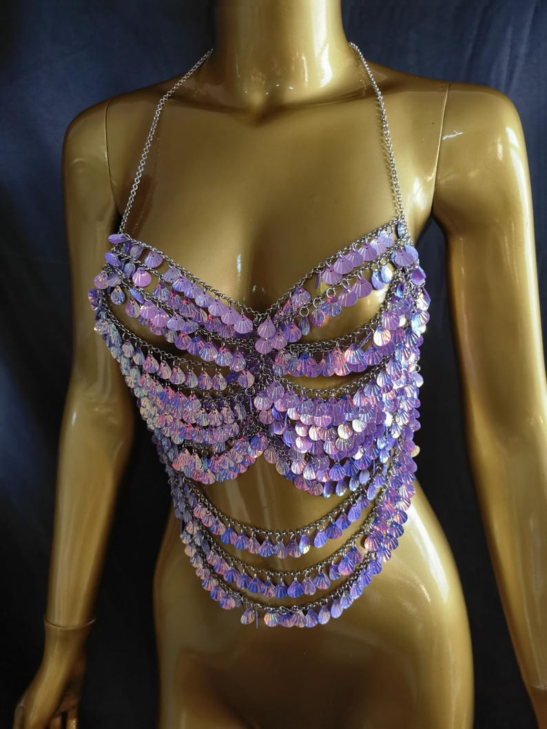 May include: A halter-style top constructed from silver-toned metal chains and iridescent, light purple, shell-shaped sequins. The garment features multiple draped layers of these shimmering, scale-like discs that create a textured, reflective surface.