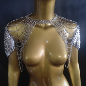 May include: Silver chainmail shoulder armour with scale-like pauldrons and a choker-style necklace. The armour is displayed on a mannequin with a gold finish, showcasing the intricate design and metallic sheen.