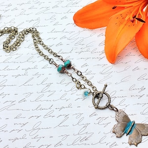 May include: A bronze butterfly pendant necklace with turquoise and copper beads. The necklace is on a white background with a lily flower.