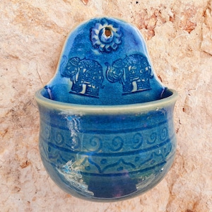 Könnte beinhalten: Eine blaue Keramik-Wandtasche mit einem dekorativen Elefanten-Design. Die Tasche hat eine abgerundete Schalenform mit einem detaillierten Muster am Rand. Die Oberseite zeigt ein Blumenmuster und ein Loch zum Aufhängen.