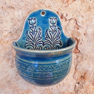 Könnte beinhalten: Eine blaue Keramik-Wandtasche mit floralem Design. Die Tasche hat eine abgerundete Schalenform mit einem dekorativen Band in der Mitte. Der obere Teil zeigt zwei stilisierte Blumenmotive. Die Tasche ist an einer strukturierten, hellbraunen Wand befestigt.