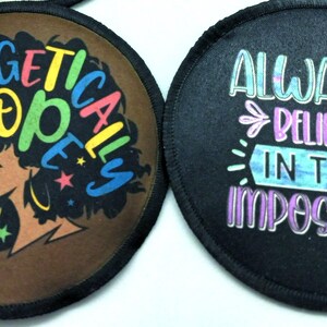 Two round black fabric patches with colorful designs. The first patch features a brown background with a silhouette of a woman with an afro hairstyle. The text "Unapologetically Dope" is written in rainbow colors around the woman's head. The second patch features a black background with the text "Always Believe in the Impossible" written in rainbow colors