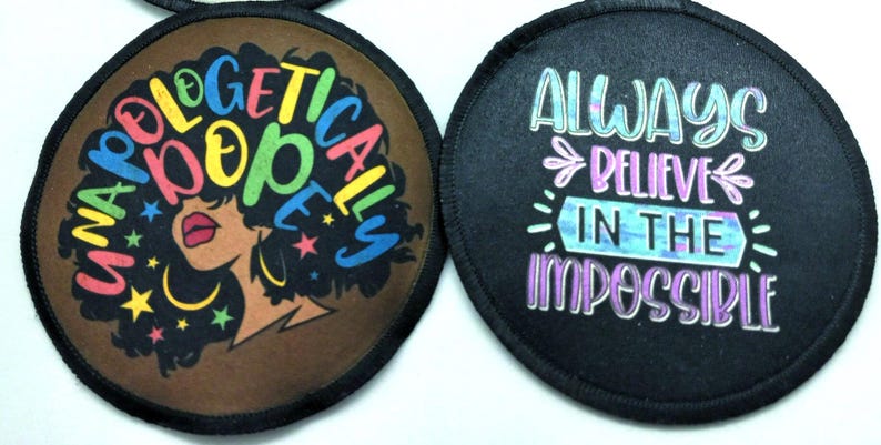 Two round black fabric patches with colorful designs. The first patch features a brown background with a silhouette of a woman with an afro hairstyle. The text "Unapologetically Dope" is written in rainbow colors around the woman's head. The second patch features a black background with the text "Always Believe in the Impossible" written in rainbow colors