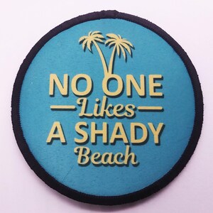 Round iron-on patch with teal background and yellow text reading “No One Likes a Shady Beach,” featuring palm tree graphic and black merrowed border.