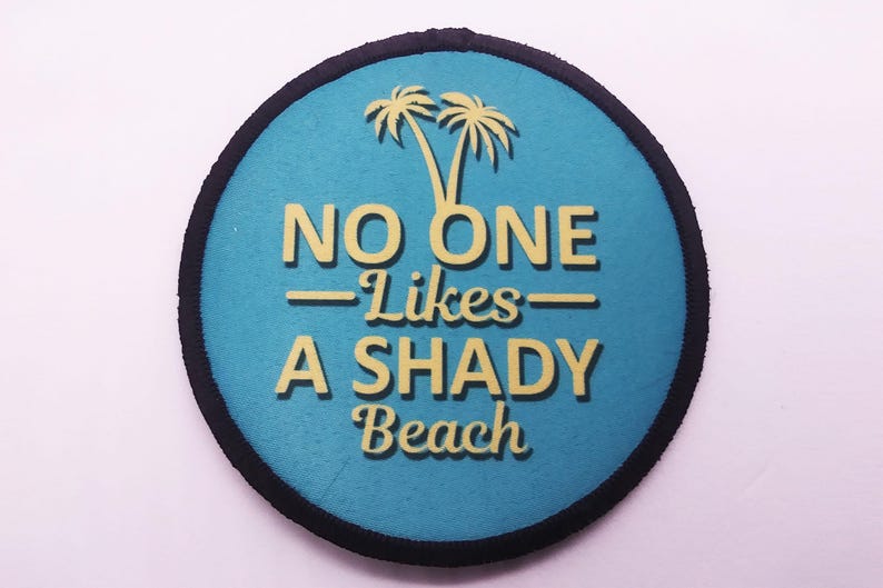 Round iron-on patch with teal background and yellow text reading “No One Likes a Shady Beach,” featuring palm tree graphic and black merrowed border.