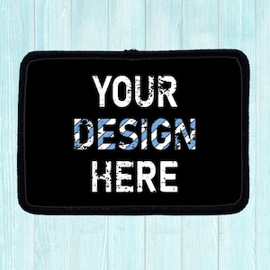 Black rectangular patch with the words "YOUR DESIGN HERE" in distressed white font. The word "DESIGN" has light blue stripes. The patch has a black border and is on a light blue wooden surface.