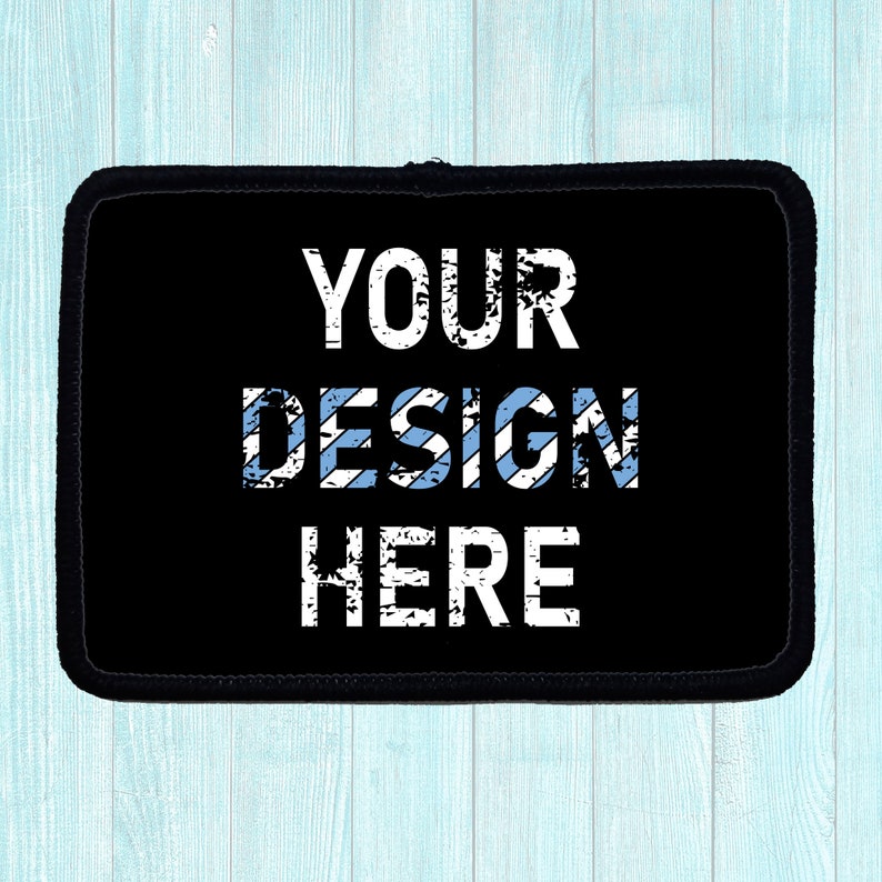 Black rectangular patch with the words "YOUR DESIGN HERE" in distressed white font. The word "DESIGN" has light blue stripes. The patch has a black border and is on a light blue wooden surface.