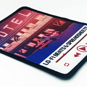 Angled side view of a full-color printed rectangle patch featuring a motel illustration and text reading “Lo-Fi Beats & Spreadsheets.”
