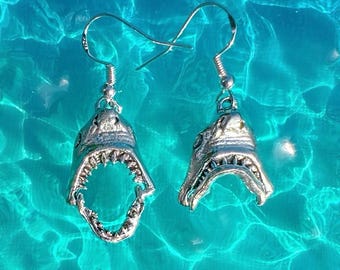May include: A pair of silver-toned shark head earrings featuring detailed snouts and open jaws with visible teeth. Each earring is attached to a standard silver-colored metal fishhook wire, designed for hanging from the earlobe with a dangling style.