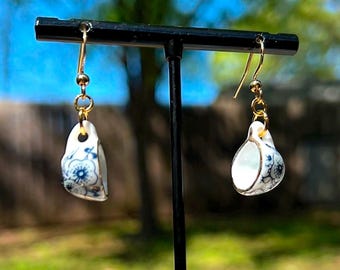 May include: A pair of gold dangle earrings with a small porcelain teacup charm. The teacups are white with blue and gold floral designs.