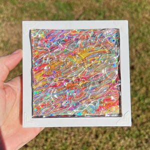 May include: A square white frame containing a piece of art with a swirling, marbled pattern of red, yellow, blue, and orange colors. The surface has a reflective, holographic finish that creates a shimmering, multi-colored effect.