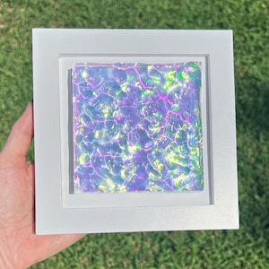 Könnte beinhalten: Ein quadratischer weißer Rahmen mit einem irisierenden Material, das eine wellige, wasserähnliche Textur aufweist. Die Oberfläche zeigt ein lebendiges Farbspektrum mit hellen violetten, rosa und gelben Reflexen, die einen schimmernden Effekt erzeugen.