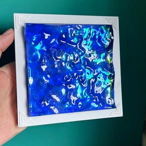May include: A square white frame containing a textured, translucent blue panel with a rippled, wavy surface. The material exhibits iridescent highlights in shades of green and silver, creating a reflective, water-like appearance within the border.