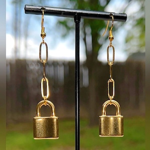Puede incluir: Un par de pendientes dorados con forma de candado. Cada pendiente presenta un pequeño colgante rectangular con forma de candado suspendido de una cadena de eslabones ovalados y un gancho. El fondo es borroso, verde y azul.