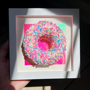 May include: A square white frame containing a three-dimensional donut sculpture covered in multi-colored sprinkles in shades of pink, blue, and yellow. The donut is centered against a vibrant, reflective, iridescent pink square background.