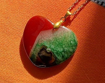 Puede incluir: Un collar con colgante en forma de corazón y cadena dorada. El colgante es una mezcla de resina roja, blanca y verde, con un patrón único. El collar se muestra sobre una superficie naranja.