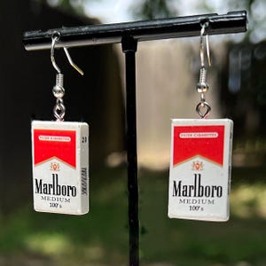 Op de afbeelding: Een paar zilveren oorhangers met miniatuur rode en witte Marlboro sigarettenpakjes bedels. De bedels hebben het Marlboro logo en de tekst "FILTER CIGARETTES", "Marlboro", "MEDIUM" en "100's".