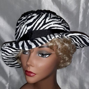 May include: A black and white zebra print hat with a black ribbon. The hat is worn by a mannequin with blonde hair.