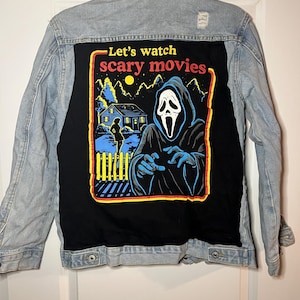 May include: Light blue denim jacket with a black back panel featuring a graphic from the movie Scream. The graphic includes the text "Let's watch scary movies" and an illustration of the Ghostface killer.