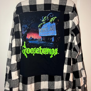 May include: Black and white plaid flannel shirt with a black graphic on the back. The graphic features the words "Welcome to Horrorland" and "Goosebumps" in green, with a monster and amusement park scene.