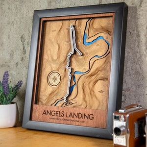 Może przedstawiać: Drewniana mapa Angels Landing w Parku Narodowym Zion w stanie Utah, umieszczona w ramie. Mapa jest wycięta laserowo i przedstawia błękitną rzekę wijącą się przez góry. Rama jest czarna z drewnianym wkładką.