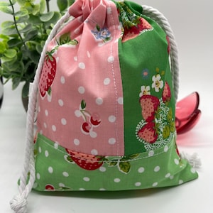 May include: A patchwork drawstring bag with a white rope closure. The bag features pink and green panels with strawberry and cherry patterns, and white polka dots. The bag is approximately 15 cm tall.
