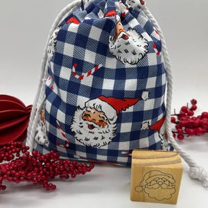 May include: A drawstring gift bag with a blue and white gingham pattern, featuring Santa Claus illustrations and candy canes. A small wooden stamp with a Santa Claus face is also present. The bag has a white rope drawstring.