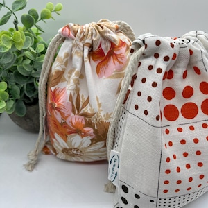 May include: Two reusable produce bags, one with a floral print and the other with a black and orange polka dot pattern. Both bags have white fabric and are made of natural materials. The bags have a drawstring closure and are labelled "Re-Wrap It All".