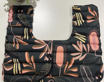 May include: A U-shaped neck wrap featuring a dark charcoal gray fabric with a repeating pattern of black cockatoos, pink banksia flowers, and muted orange foliage. The wrap is constructed with horizontal quilted channels throughout the design.