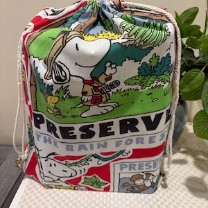 May include: A drawstring bag with a Snoopy and Woodstock rainforest design. The bag is white, red, and green, with the words "Preserve the Rainforest" printed on it. It has a white drawstring closure and is made of fabric.