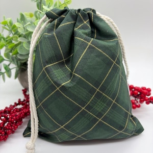 May include: A dark green fabric drawstring pouch featuring a gold-coloured plaid pattern. The bag is cinched at the top with thick, off-white twisted cotton rope cords that are knotted at the ends, creating a gathered closure for the pouch.
