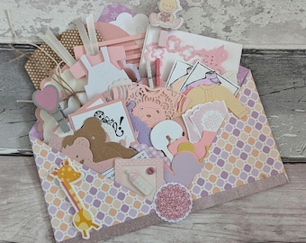 Könnte beinhalten: Ein offener Umschlag gefüllt mit Baby-Themen-Papier-Ausschnitten und Verzierungen. Der Inhalt umfasst Kleiderformen, Herzen, eine Giraffe, eine Babyflasche und einen Teddybären. Der Umschlag hat ein rosa-pfirsichfarbenes geometrisches Muster mit einem glitzernden Rand.