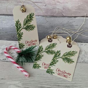 May include: Cream-colored gift tags with green fir branch designs and the words "Christmas - GREETINGS" in red. Each tag has a small gold bell attached. A red and white candy cane and greenery are also present.