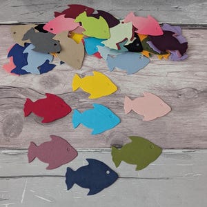Puede incluir: Una colección de peces de fieltro coloridos en varios tonos de azul, verde, amarillo, rosa, rojo y morado. Los peces están esparcidos sobre una superficie clara con vetas de madera, creando un arreglo decorativo.