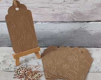 Könnte beinhalten: Braune Geschenkpapieranhänger mit gewelltem Rand und geprägten Wintermotiven, darunter Schneeflocken und Rentiere. Ein Anhänger wird auf einer kleinen Holzstaffelei präsentiert, mit einem Stapel Anhänger und Holzschneeflocken in der Nähe.