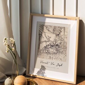 May include: A framed black and white line drawing of a cafe terrace at night by Vincent Van Gogh. The drawing is in a light wood frame and is sitting on a wooden floor.