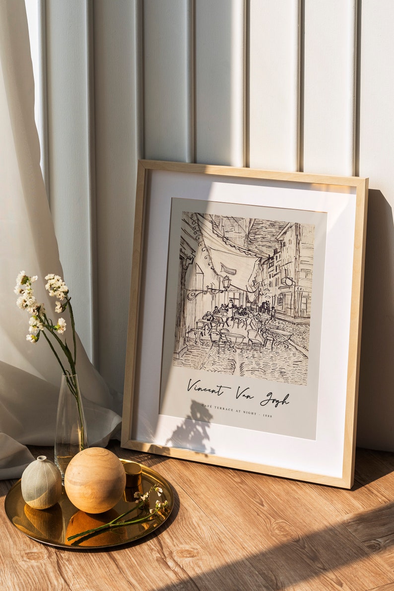 May include: A framed black and white line drawing of a cafe terrace at night by Vincent Van Gogh. The drawing is in a light wood frame and is sitting on a wooden floor.